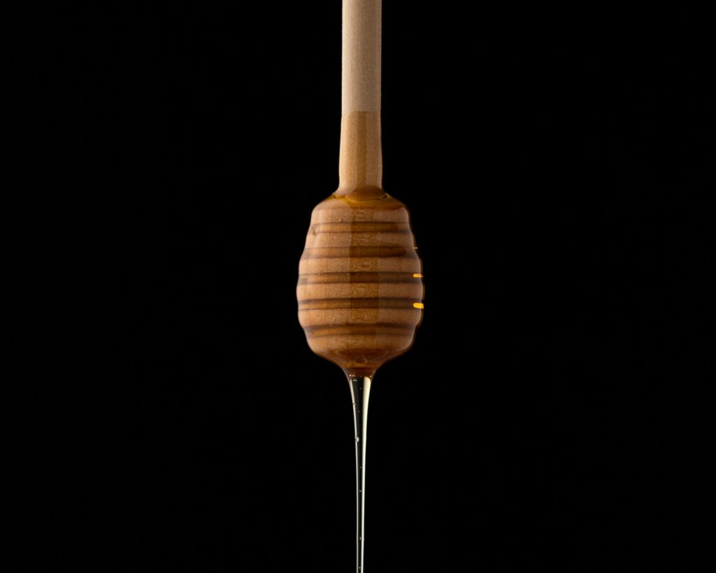 A long, slow  drip of honey coming off of a wooden honey comb, over a black background.