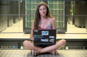A woman with long brown wavy HAIR sits cross-legged on the floor in front of a rack of servers - part of Maranostru 4. She has a black laptop with stickers on the lid in her lap. The stickers read "pop", "mont blanc", "OpenMP" and "HPC Bottleneck Chasers". She's wearing a colorful paisley dress and sandals.