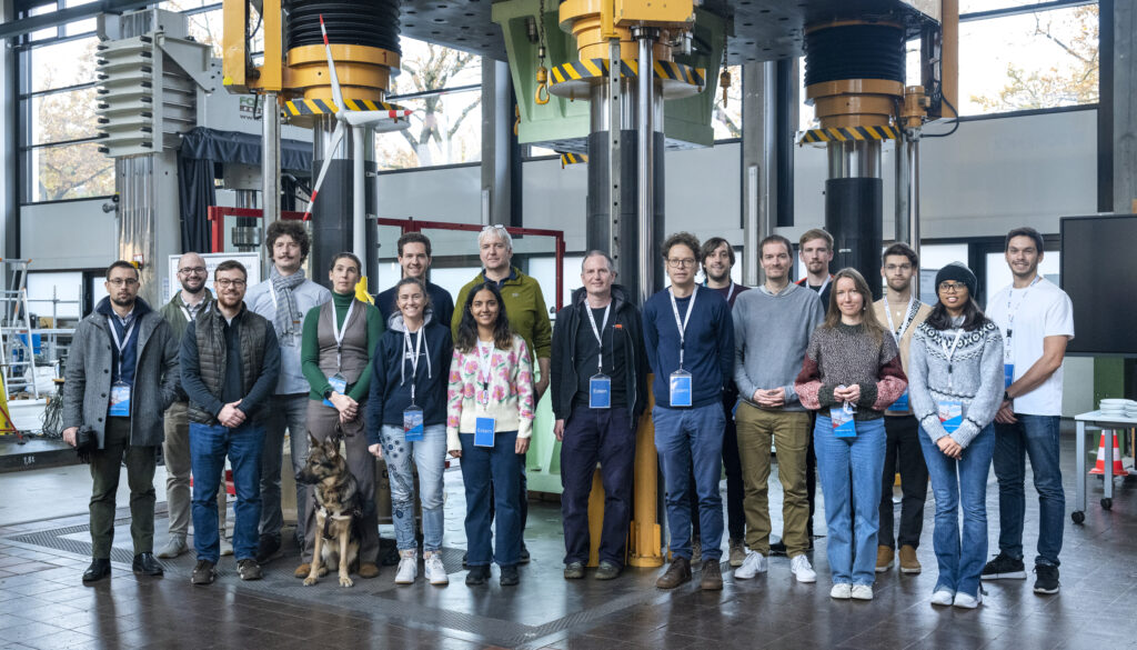 A group of 17 people is standing in a test facility during an all hands meeting for the Center of Excellence in Exascale CFD, hosted at the Bundesanstalt für Materialforschung und -prüfung (BAM) in November 2024. The group includes both men and women, all smiling and facing the camera, with some wearing lanyards and badges. A German Shepherd guide dog is sitting in front of the group next to her handler. The setting is an industrial space with large machinery in the background, featuring vertical structures with yellow and black striped markings. The facility has high ceilings and large windows.