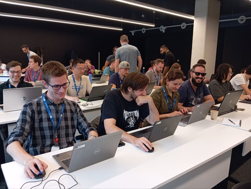 A group of people are seated in a classroom setting at the MNHACK hackathon held at the Barcelona Supercomputing Centre. They are working on laptops, with two rows of tables filled with participants. Most individuals are wearing blue lanyards with name badges, indicating their participation in the event. The atmosphere is lively and collaborative, with several people smiling and interacting with each other. The room has a modern design, featuring black walls and ceiling, and linear lighting fixtures.