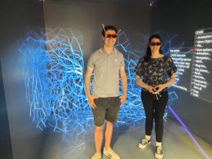 Two people stand in a virtual reality environment at the HLRS cave, engaging with a Vistle visualization. Both are wearing 3D glasses and holding white controllers. The man on the left is in a light gray polo shirt and dark shorts, while the woman on the right wears a dark floral blouse and black pants. The background displays blue geometric wireframe patterns and a menu with options like "View options" and "Scale" on the right, indicating an interactive digital setup.
