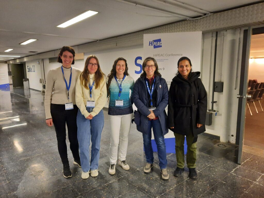 Five women standing in a hallway, each wearing a conference badge with a blue lanyard. From left to right: 1. Susanne Malheiros has short brown hair and is wearing a beige turtleneck sweater with black pants and black shoes. 2. Anna Schwarz has long straight brown hair and glasses, wearing a cream-colored sweater, light blue jeans, and white sneakers. 3. 
Marta Garcia has long curly brown hair, wearing a blue and white gradient sweater, white pants, and beige sneakers. 4. Rosa Badia has short brown hair and glasses, wearing a blue jacket over a maroon top, blue jeans, and gray sneakers. 5. Pratibha Raghupati has long black hair tied back, wearing a black coat, olive green pants, and black shoes. They are standing in front of a sign for the HiPEAC Conference, with a polished floor and fluorescent lighting in the hallway.