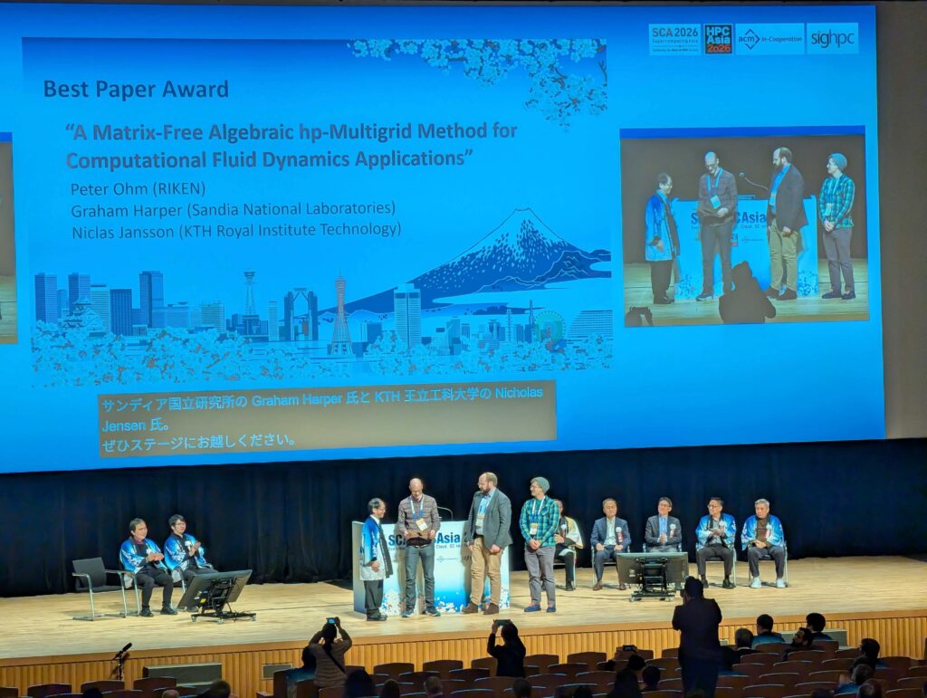 Conference hall with a large screen displaying a slide titled “Best Paper Award” for a paper called “A Matrix-Free Algebraic hp-Multigrid Method for Computational Fluid Dynamics Applications,” listing authors and recipients Peter Ohm (RIKEN), Graham Harper (Sandia National Laboratories), and Niclas Jansson (KTH Royal Institute Technology), with blue cityscape and mountain graphics and several event logos at the top right. Onstage below the screen, three people in blue happi-style jackets sit to the left, while at center a person in a blue jacket presents an award to the three listed authors standing side by side. To the right, five suited officials, some also in blue jackets, are seated in a row. In the foreground, audience members sit in theater-style seats while a few people near the front photograph the award presentation.